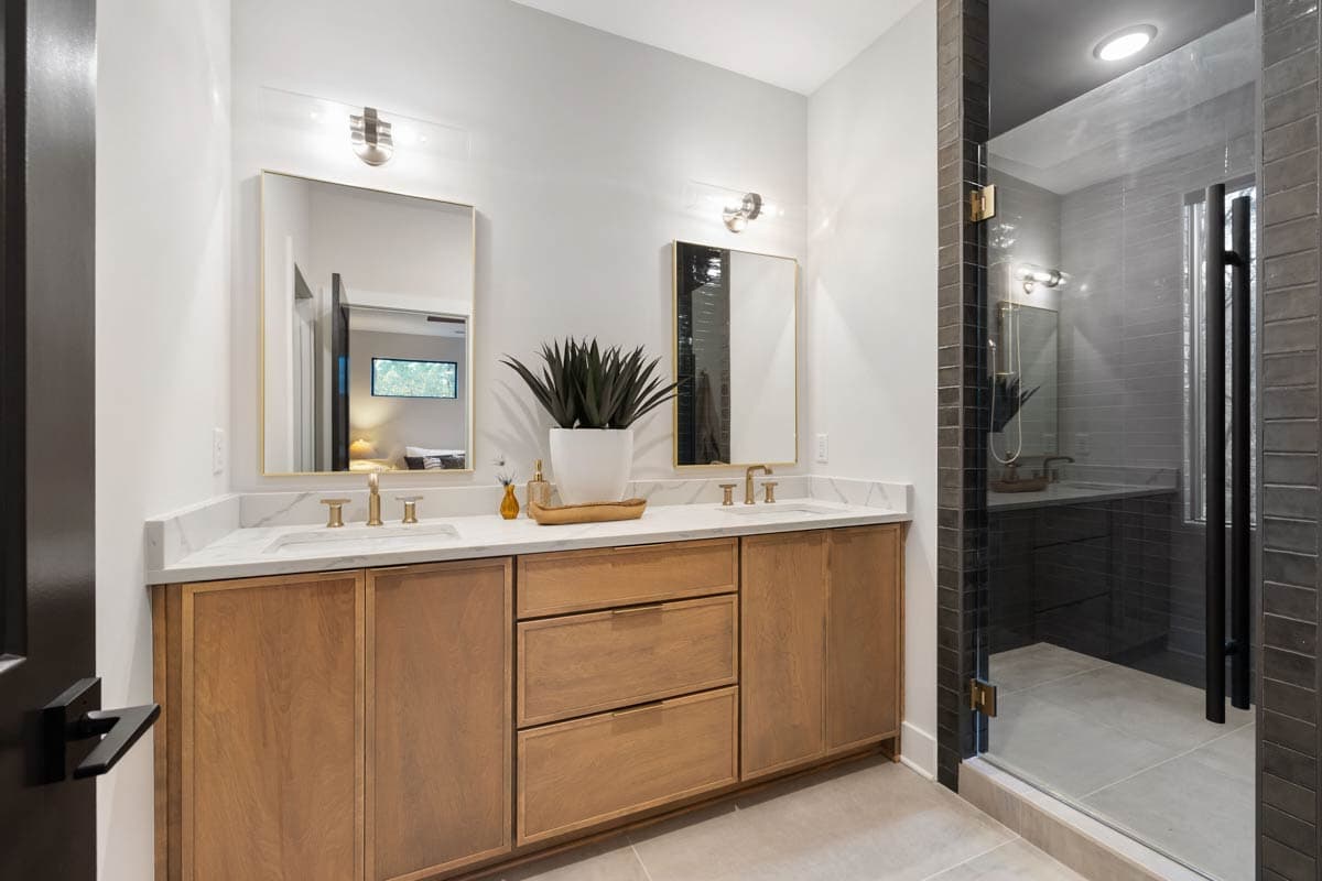 A modern bathroom features two mirrors with gold frames above wooden cabinets and a marble countertop. A large plant sits between the mirrors.
