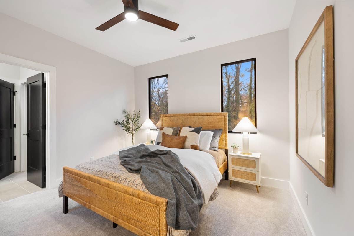 A modern bedroom features a woven headboard and bed frame. Two windows show a view of trees. White bedside tables flank the bed with lamps.