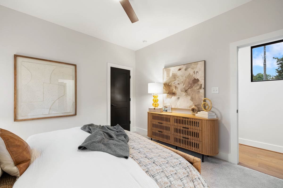 A modern bedroom features a large abstract painting above a wooden dresser. A bed with a grey throw and a framed abstract art piece complete the room.