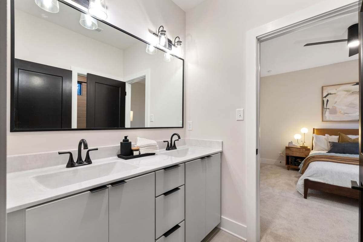 Modern bathroom with double vanity, large mirror, and open doorway to bedroom. Vanity has black fixtures, a soap dispenser, and folded towels.