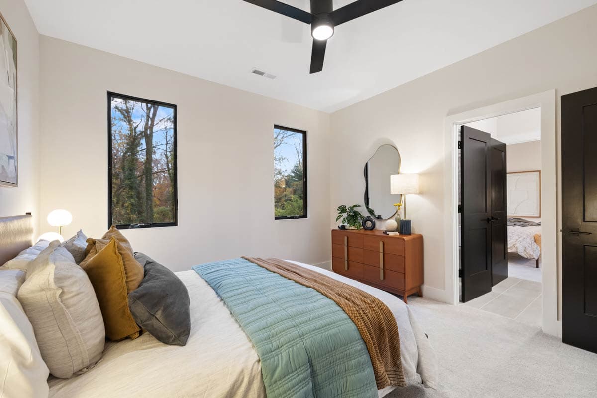 A modern bedroom features a bed with pillows and a blue quilt. A wooden dresser with a mirror sits near the windows with a view of trees.