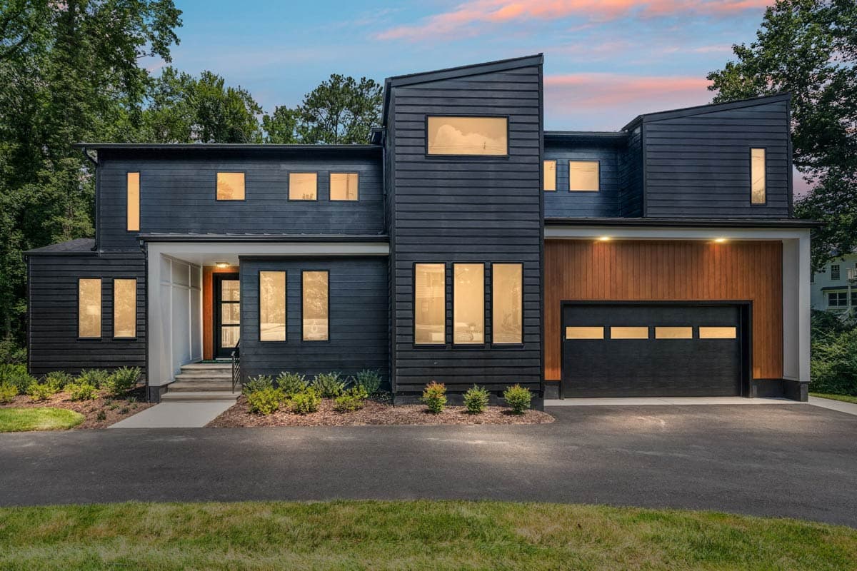 Modern two-story house with dark siding, accented by wooden paneling and large rectangular windows. A paved driveway leads to the garage.