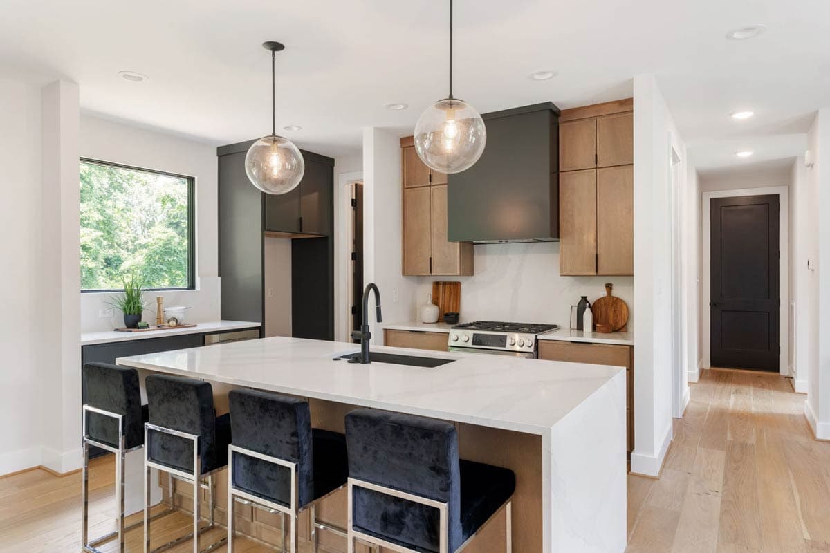 Modern kitchen with a large island, black velvet bar stools, pendant lights, and light wood cabinets. A black door is visible in the hallway.