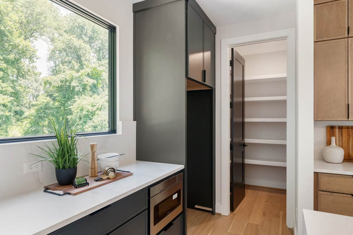 Modern kitchen with a window overlooking trees. Features include a countertop with accessories, dark cabinets, a pantry, and hardwood floors.