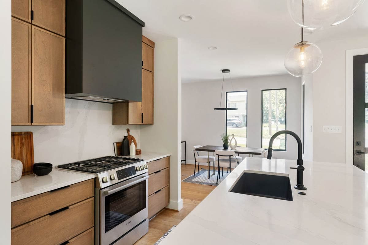 Modern kitchen with wood cabinets, stainless steel appliances, and a marble countertop. A black range hood contrasts the light space, leading to a dining area.