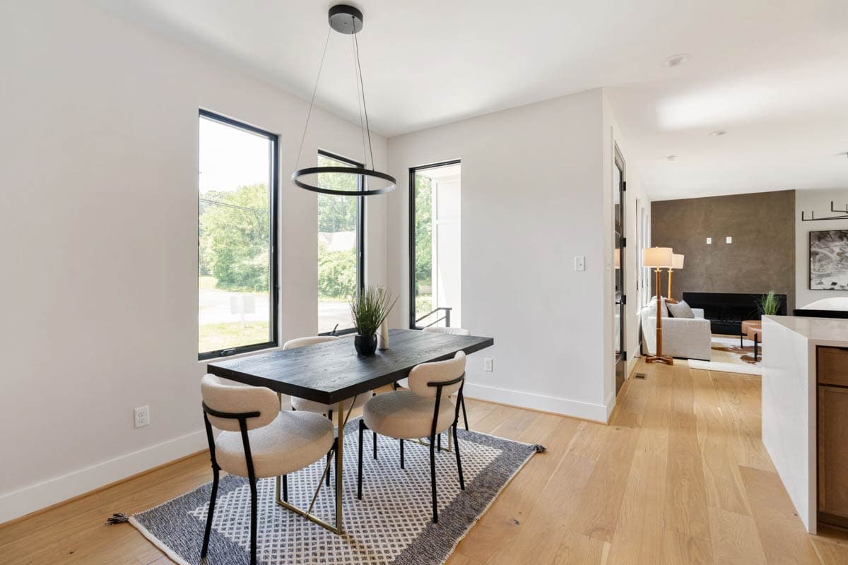 A modern dining room features a dark wood table with four beige chairs, set on a patterned rug. Large windows provide natural light.