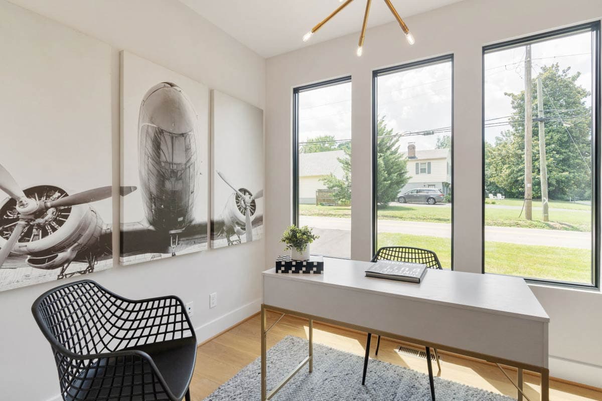 A modern home office with large windows overlooking a tree-lined street. Black and white airplane art hangs above a minimalist desk and chair.