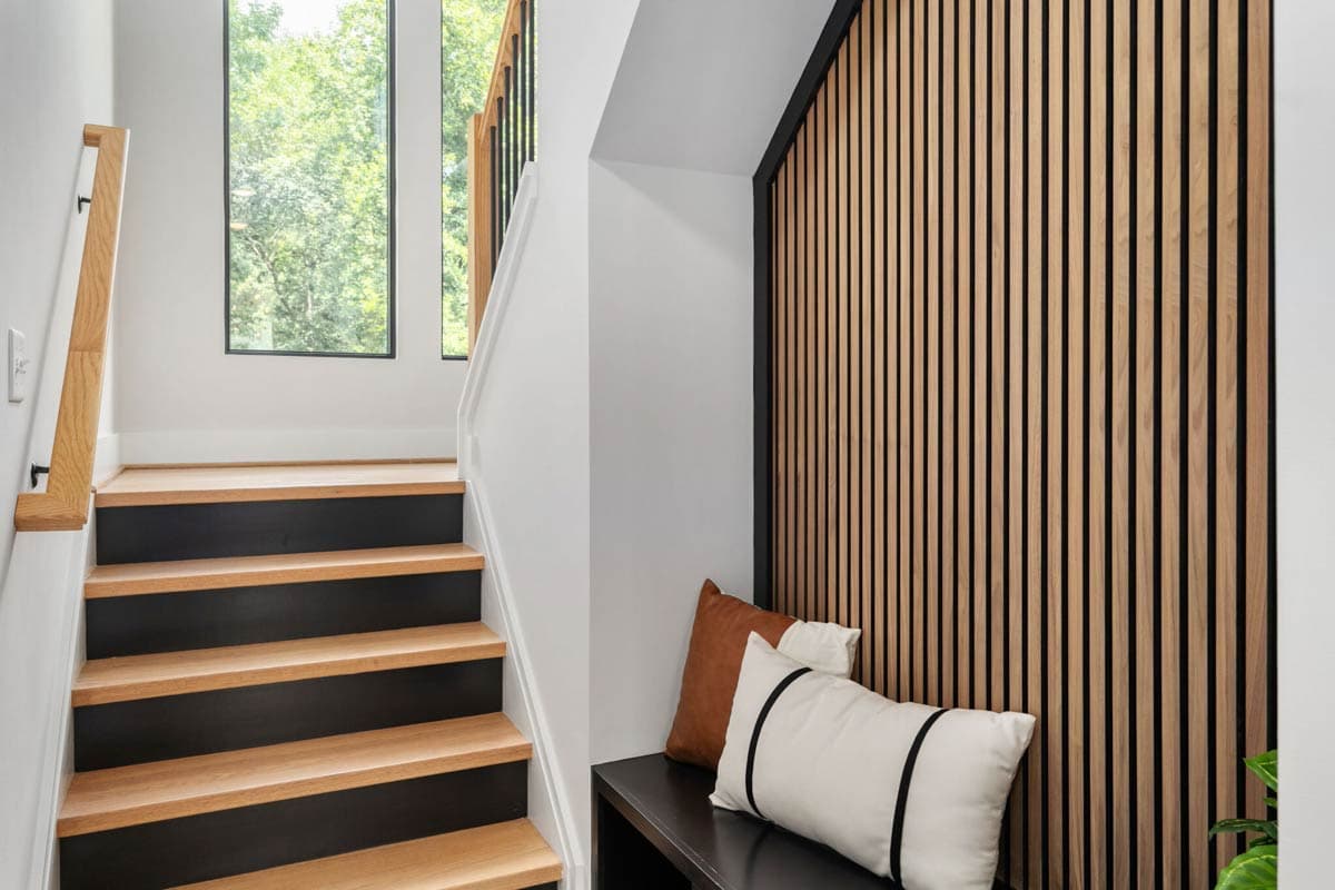 Modern interior features a staircase with wood steps, a wooden accent wall, and a bench with pillows. Windows reveal a lush green view.