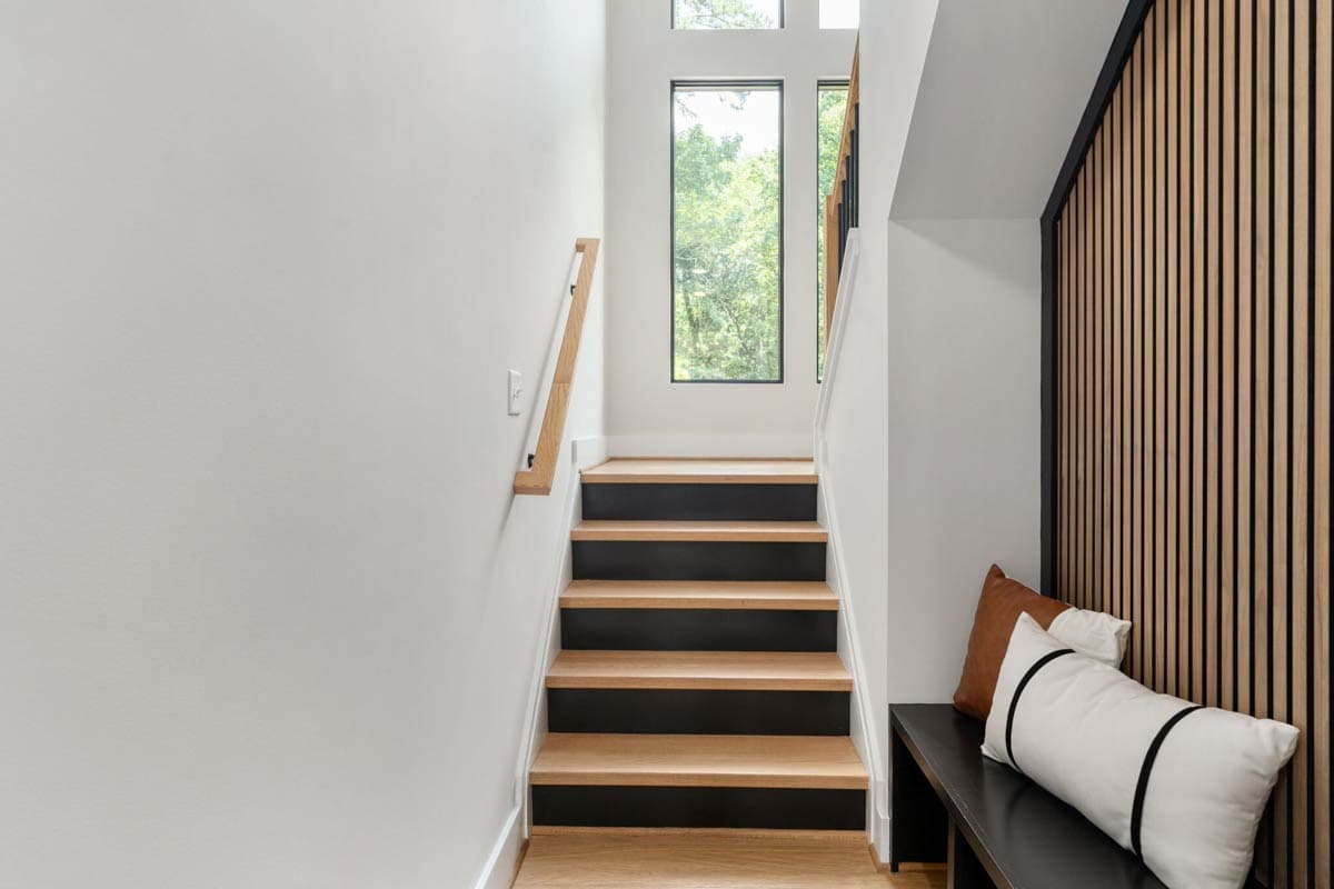 A modern interior featuring wooden stairs with black risers leading up to a window overlooking trees. A bench with pillows sits at the landing.