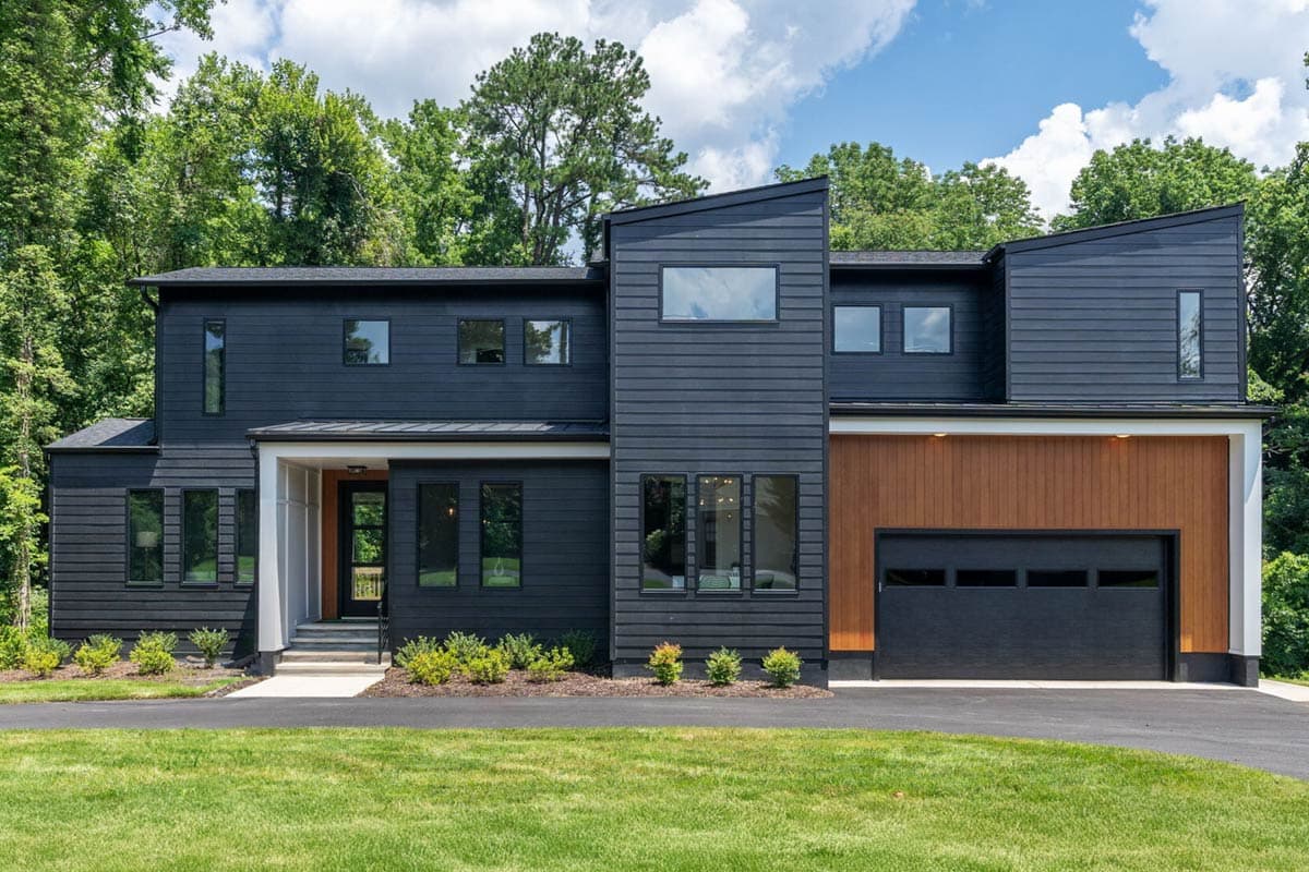 Modern two-story house with dark gray siding, accented with wood panels on the right side. Black garage door and lush green lawn in the front.