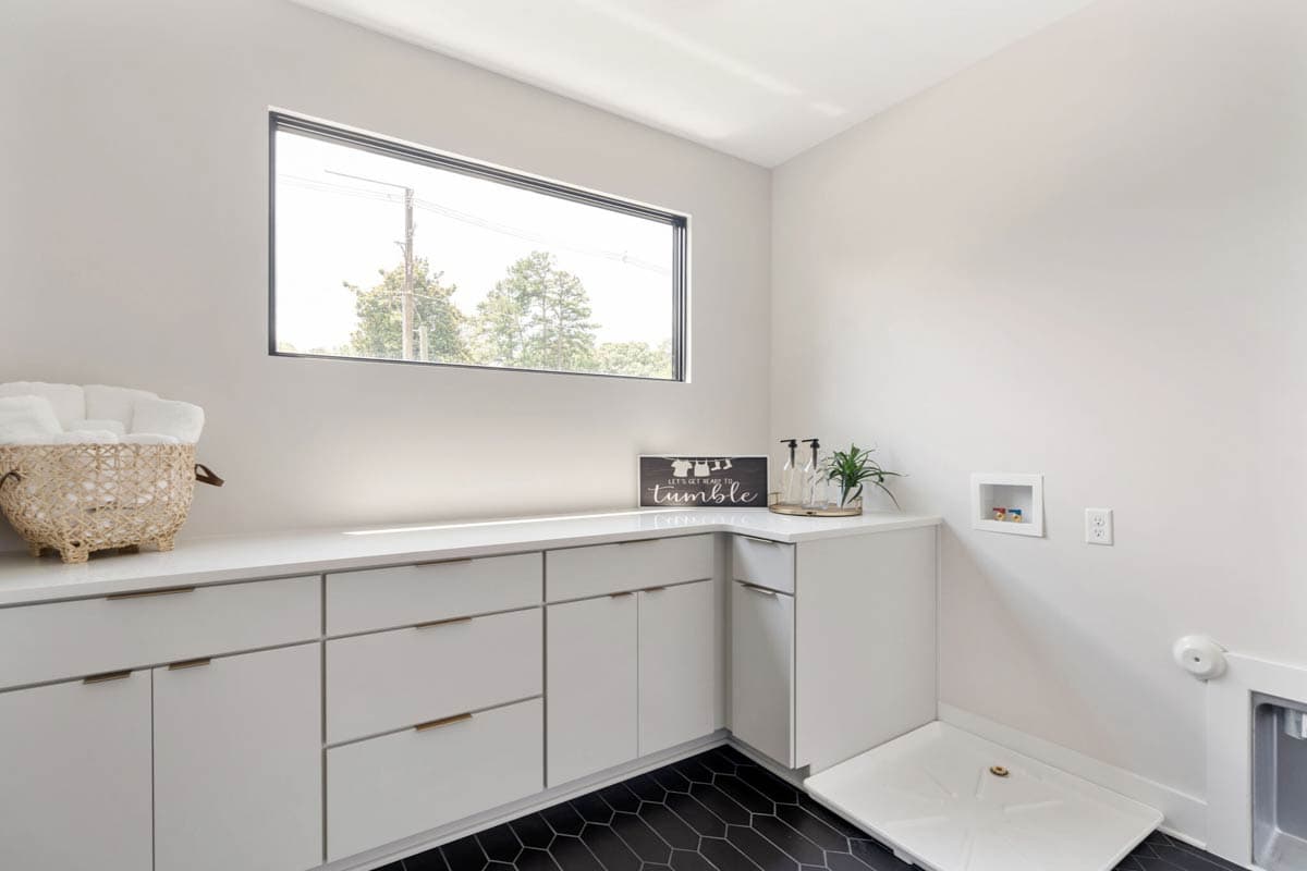 A modern laundry room with white cabinets, black tile floors, and a large window. A basket of towels sits on the counter.