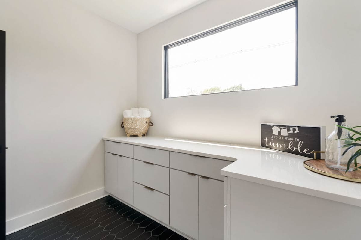 A modern laundry room with white cabinetry and countertop. A woven basket with towels, a decorative sign that reads "Let's get ready to tumble," and a large window.