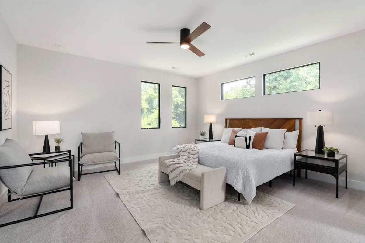 Modern bedroom with a bed, bench, and two armchairs. Large windows frame a view of trees outside.  Neutral tones dominate the space.