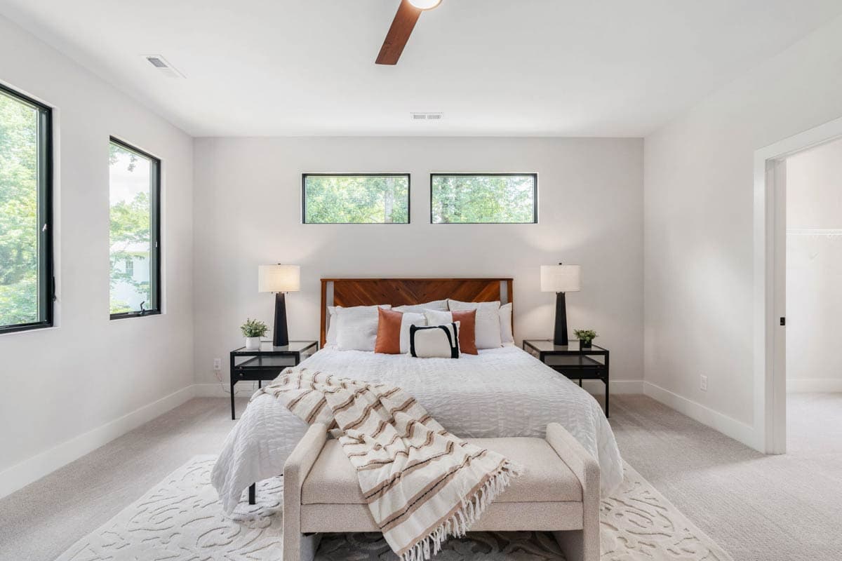 A modern bedroom with a large bed, wooden headboard, and two bedside tables with lamps. A decorative bench sits at the foot of the bed.