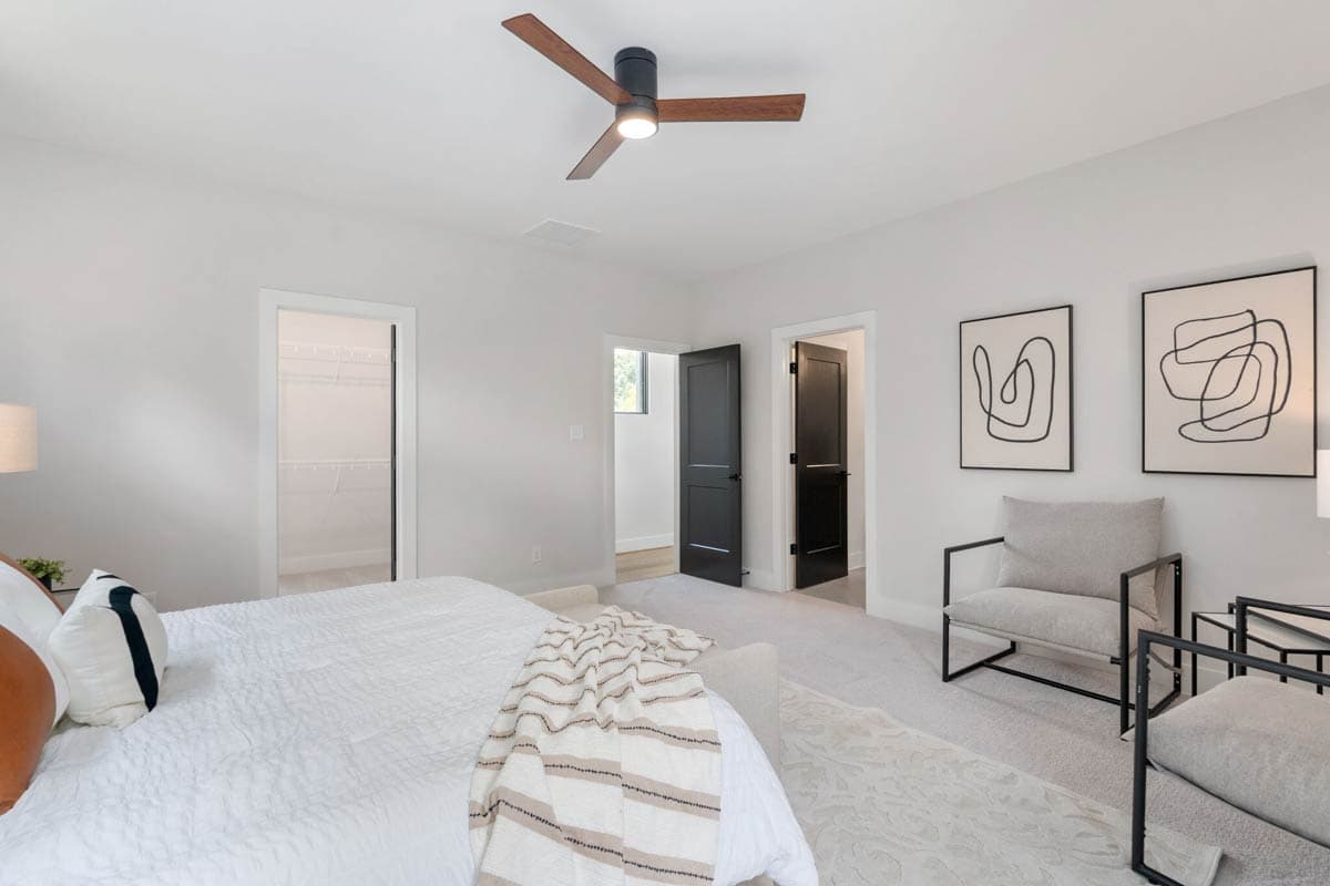 Modern bedroom with a bed, two black framed abstract art pieces, two chairs, and a ceiling fan, with two dark doors visible.