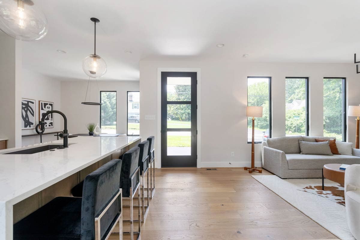 Modern, bright open-plan living space with kitchen island and black velvet bar stools, leading to a living area with a sofa and windows.