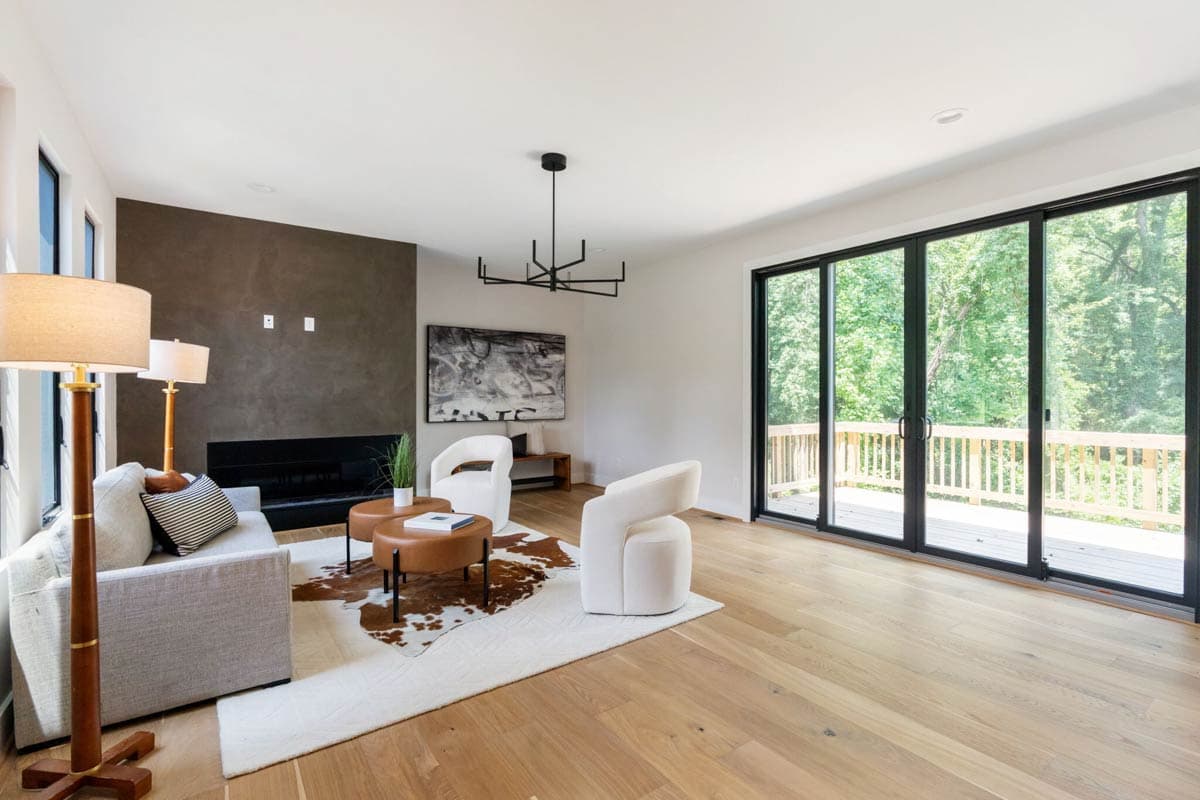 A modern living room with a fireplace, stylish furniture, and large glass doors leading to a wooden deck and a lush green forest.