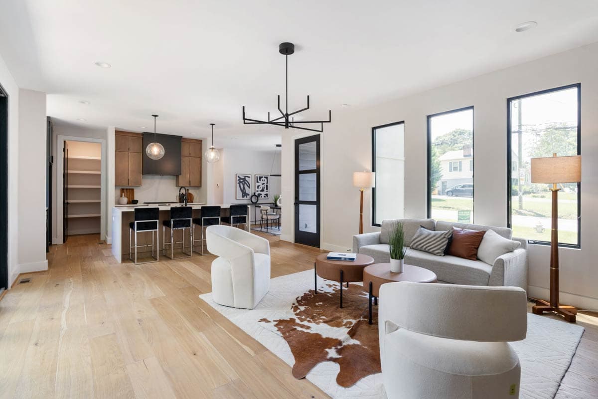 Spacious, modern living room with open kitchen, light wood floors, and a decorative cowhide rug. Two white armchairs and a grey sofa.