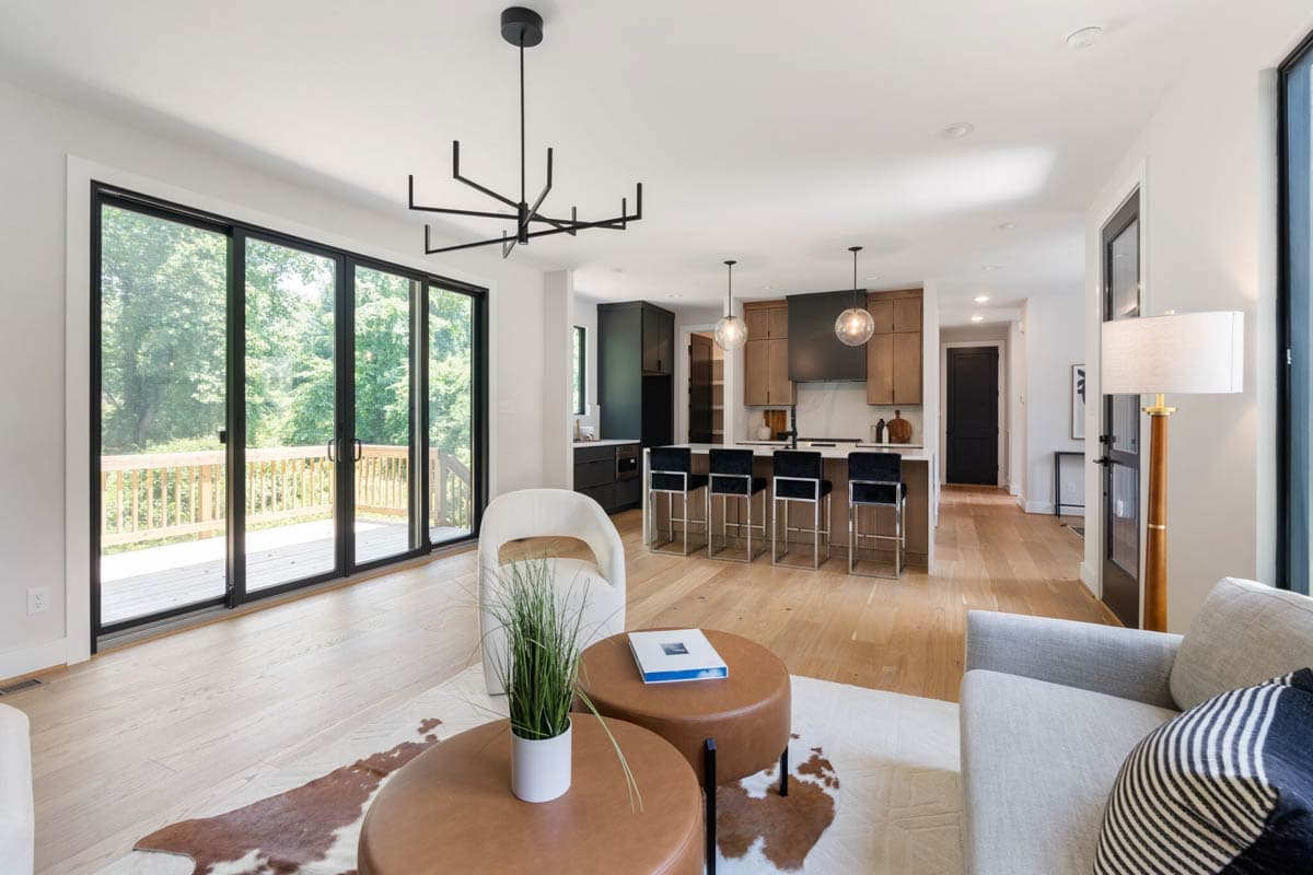 Modern, open-concept living space with a kitchen bar, light wood floors, and floor-to-ceiling glass doors leading to a deck and backyard.