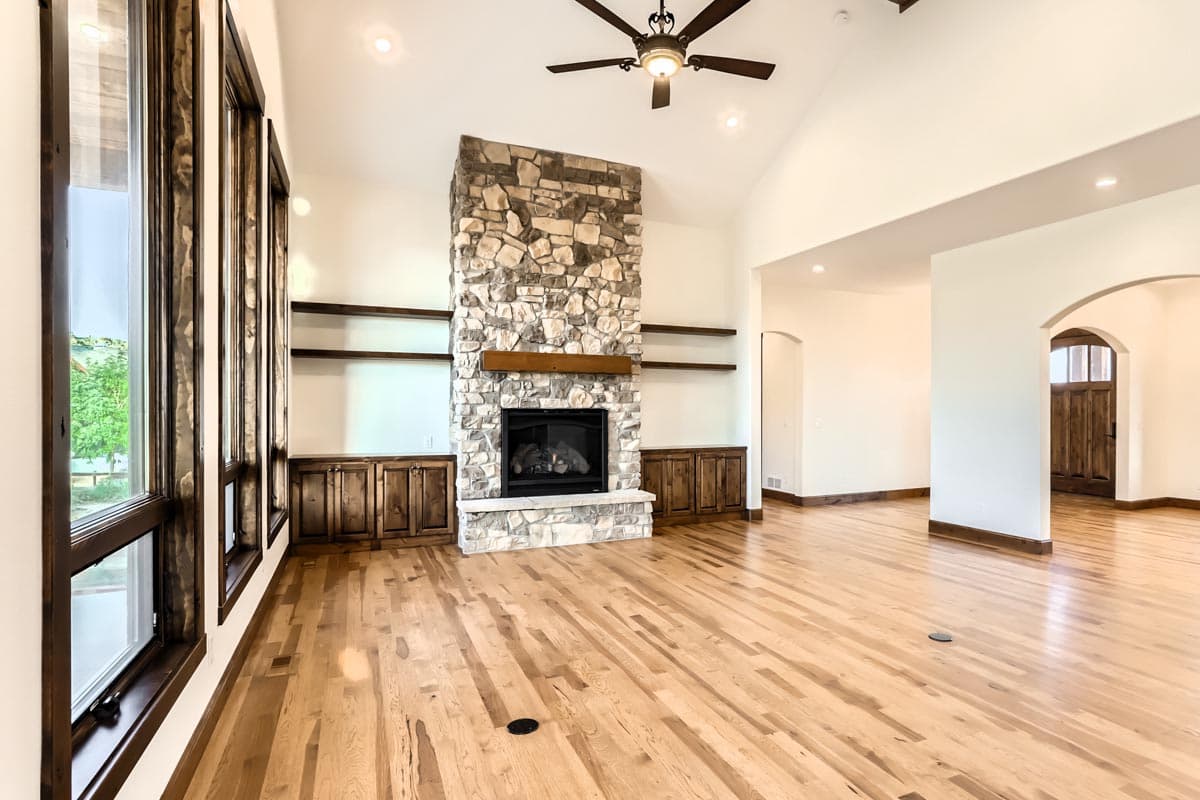 Interior view of living room with stone fireplace, vaulted ceiling, wood floors, built-in cabinets, and large windows.