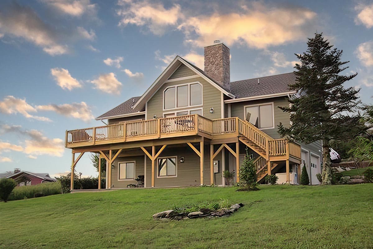 House plan exterior. Two-story house with a large wooden deck, gabled roof, and stone chimney. Garage is attached.