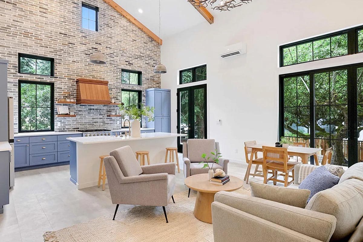 Open-concept living area with vaulted ceiling, brick accent wall, kitchen island, and large windows.