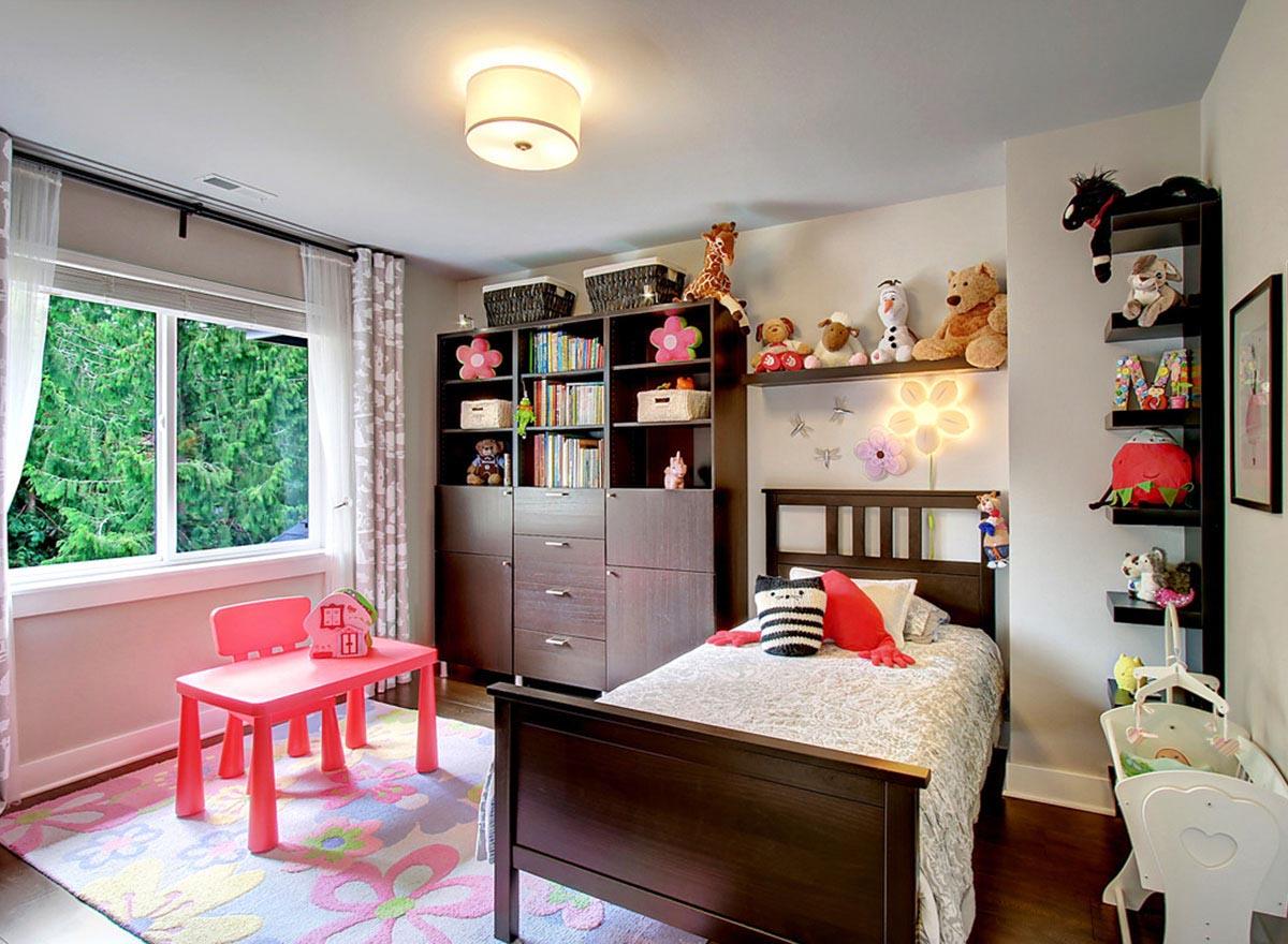 Children's bedroom with twin bed, toy storage shelves, window with curtains, and a pink play table and chairs.