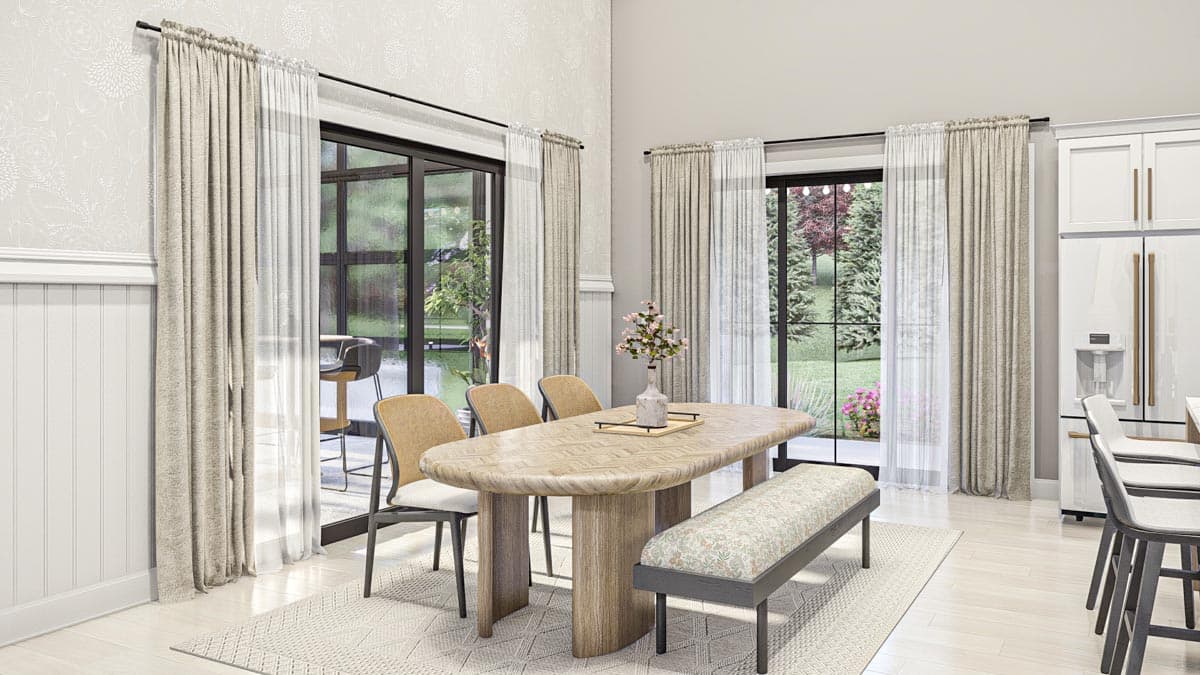 Dining area with oval table, bench, chairs, and large windows overlooking a garden. Adjacent kitchen with white cabinets and refrigerator.