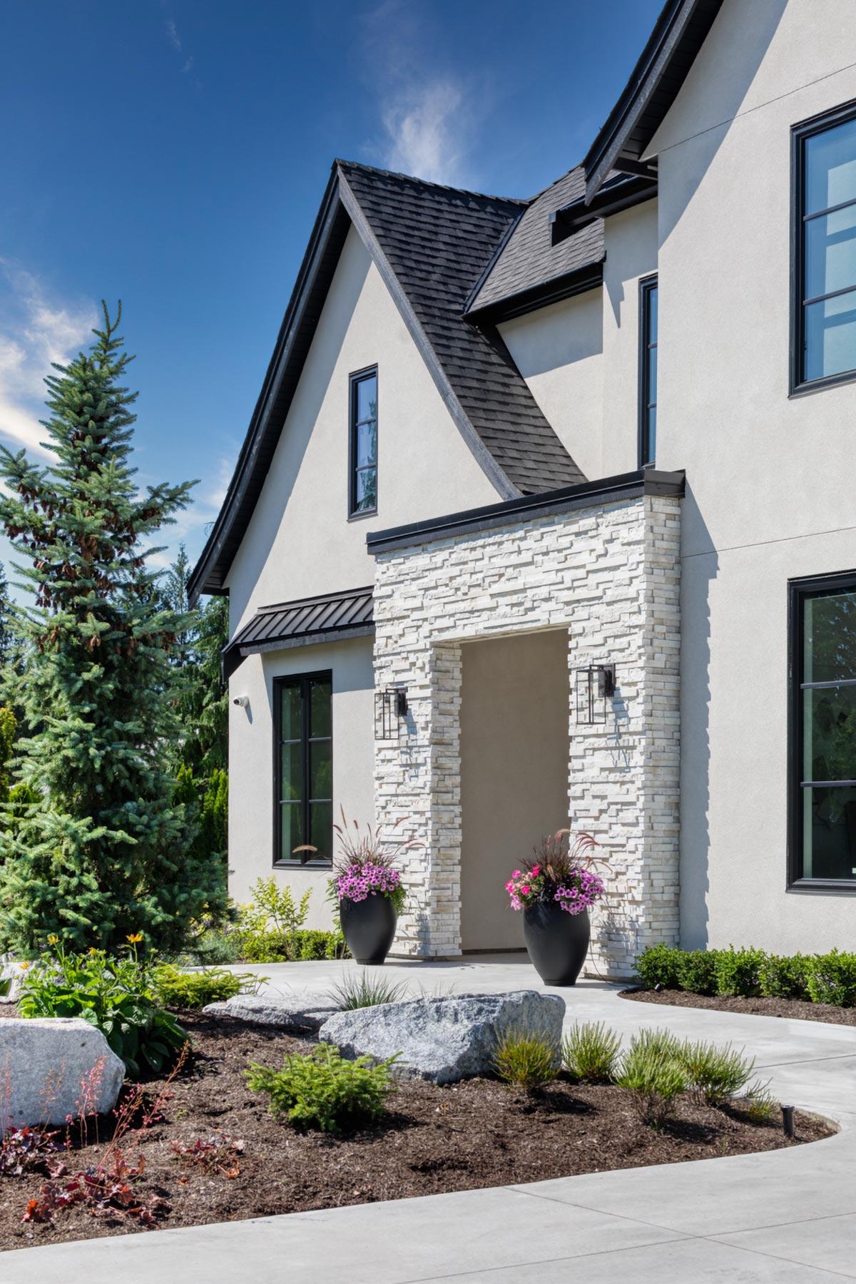House plan exterior with stone-clad entrance, stucco walls, steeply pitched gables, and dark metal roof.