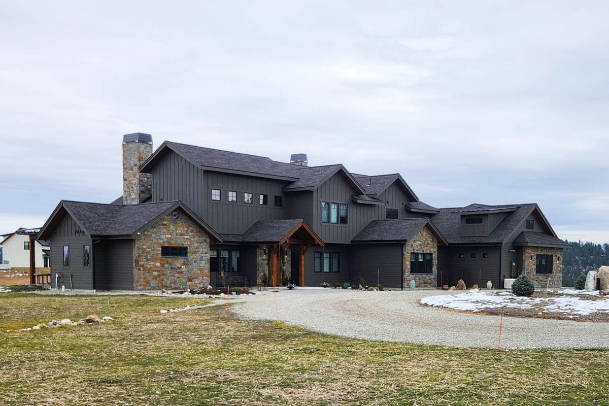 A large, modern home with a dark exterior, stone accents, and a long gravel driveway, set against a cloudy sky.