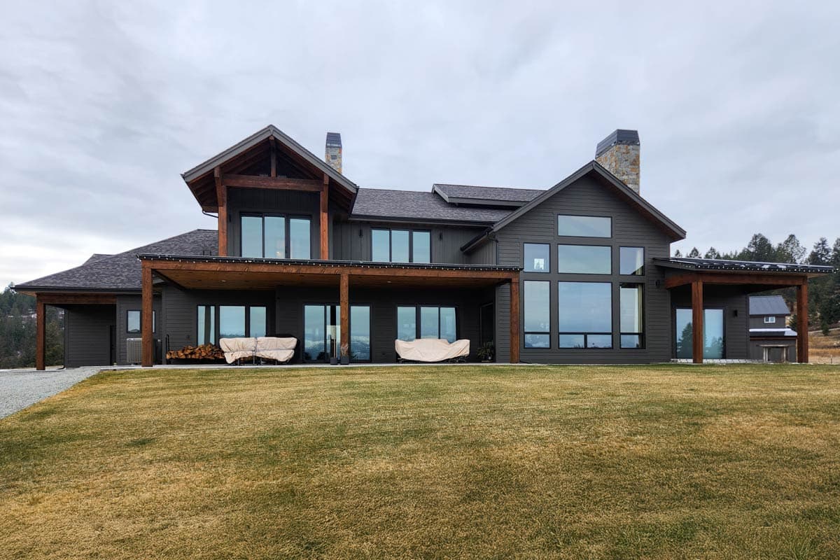 A large, modern home with dark gray siding, wooden accents, and tall windows. Two covered patios with cushioned furniture are visible.