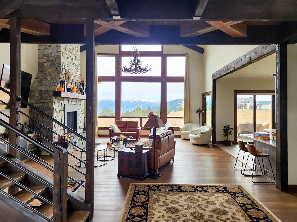 Spacious living room with large windows overlooking a mountain range. Features a stone fireplace, leather sofas, and a detailed rug.