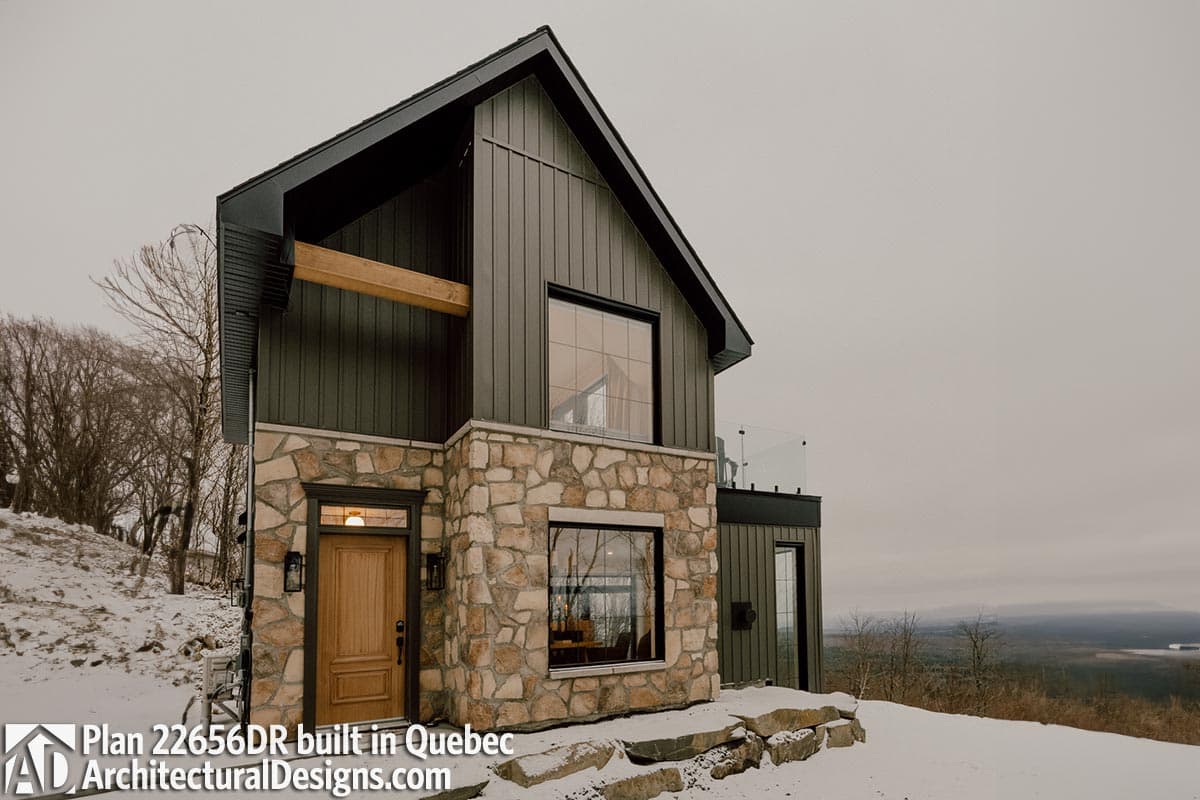 House plan exterior: Modern style with stone base, vertical siding, and exposed wood beam. Features a prominent gable roof and glass balcony.