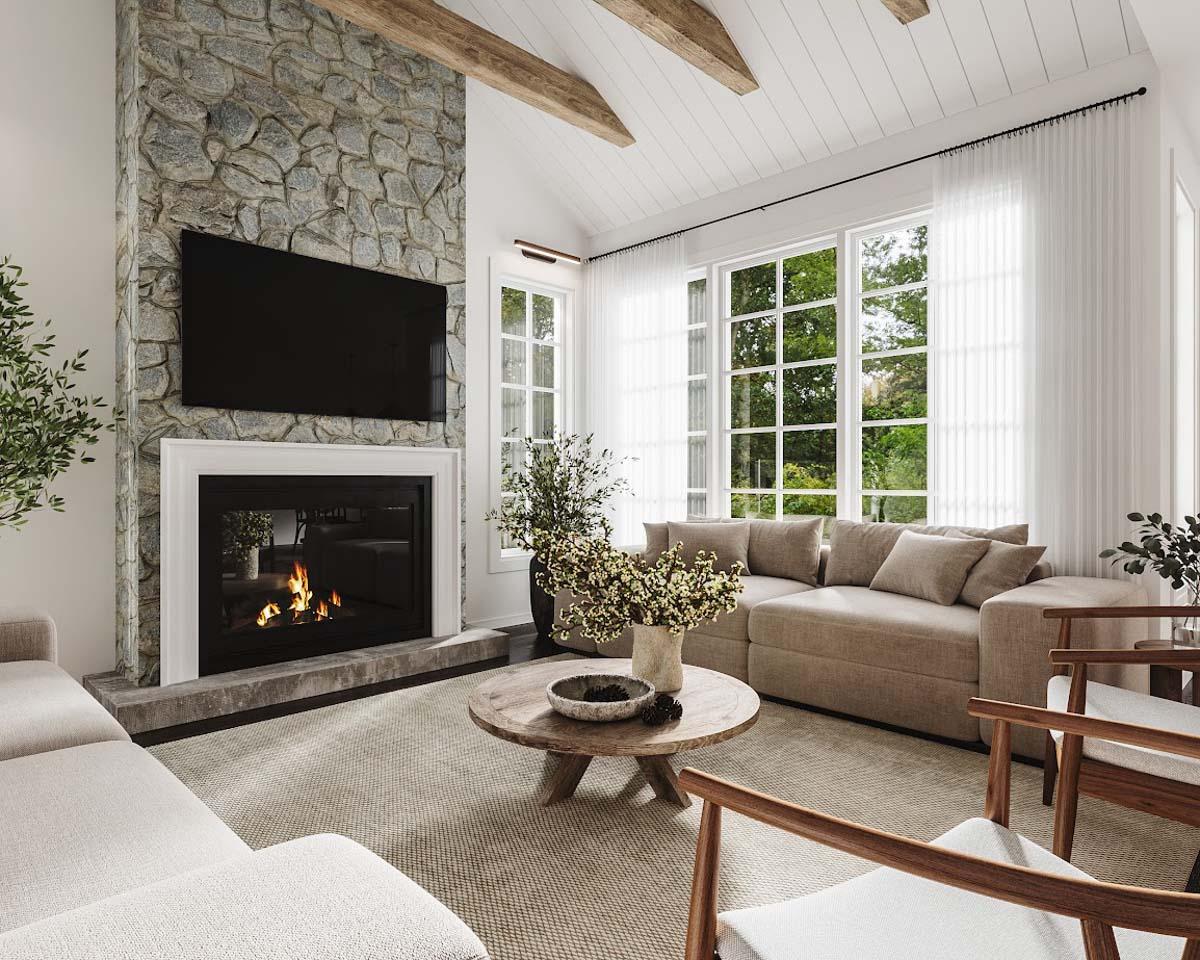 Cozy living room with stone fireplace, lit fire, and large windows looking out to greenery. A neutral-toned sofa and wooden chairs complete the scene.