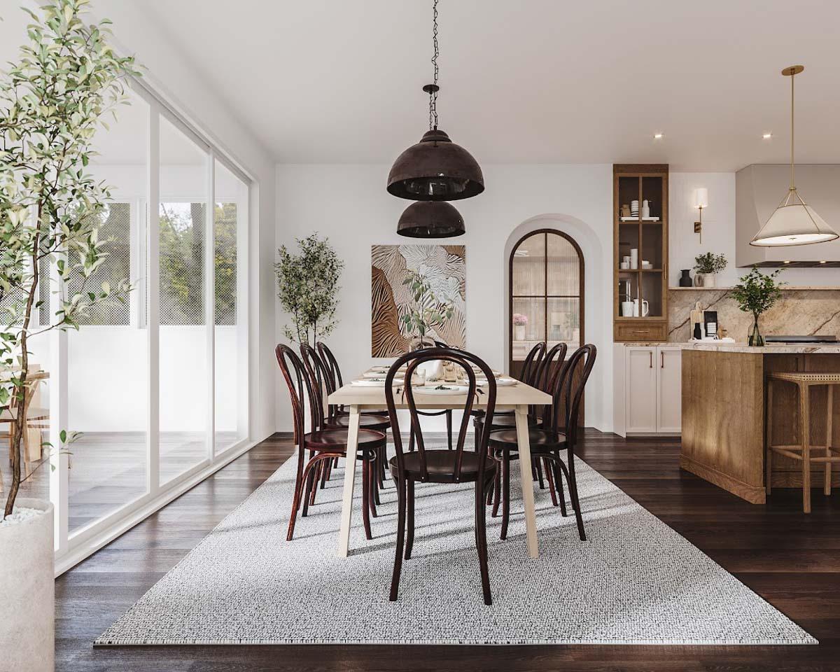 A dining room featuring a long table set for guests, wooden chairs, and decorative pendant lights. Large windows offer a view of greenery.
