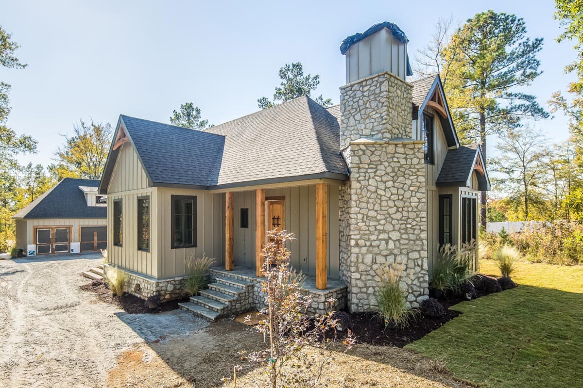 House plan exterior: Modern Farmhouse style, one-story, featuring a stone chimney, covered porch with timber posts, and a detached two-car garage.