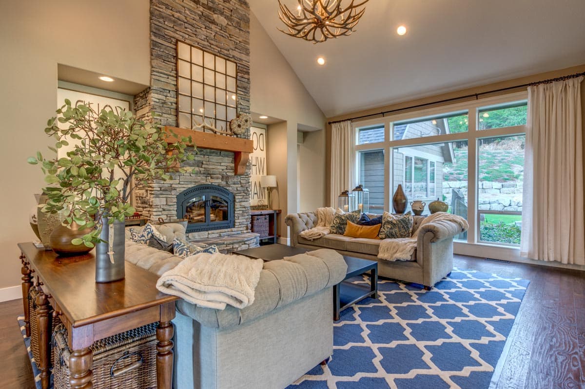 Living room with stone fireplace, vaulted ceiling, large windows, and blue geometric rug.