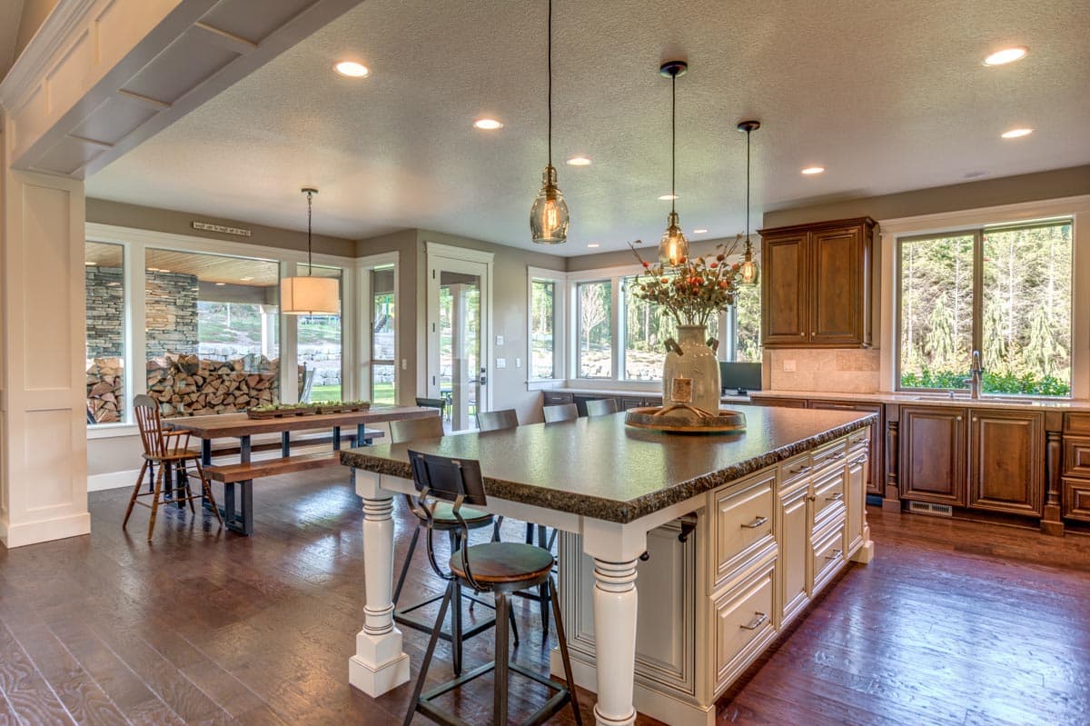 Open concept kitchen with large island, dining area with built-in bench, and extensive windows overlooking wooded landscape.