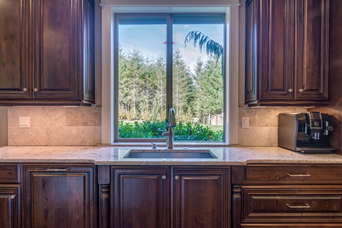 Kitchen sink centered under large window, flanked by dark wood cabinets and granite countertops. Outdoors shows trees and a small shed.