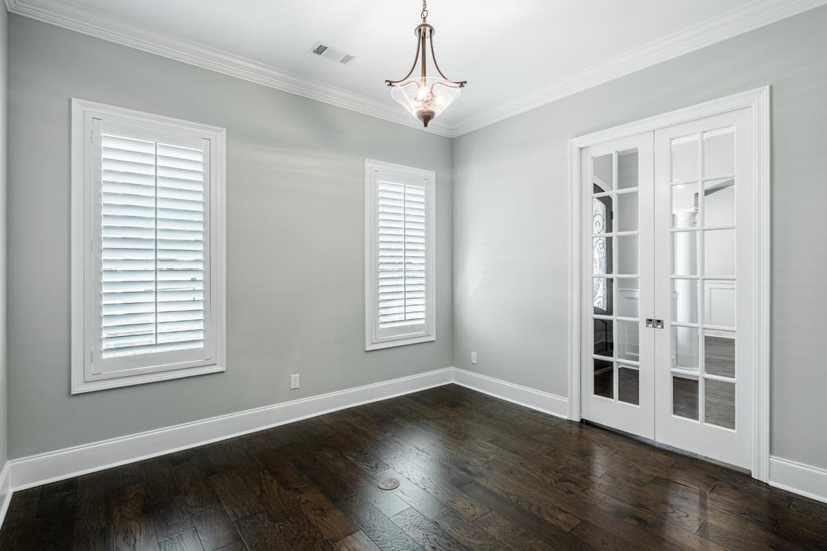 Interior view of room with double glass doors, two tall windows with shutters, and dark hardwood floors.