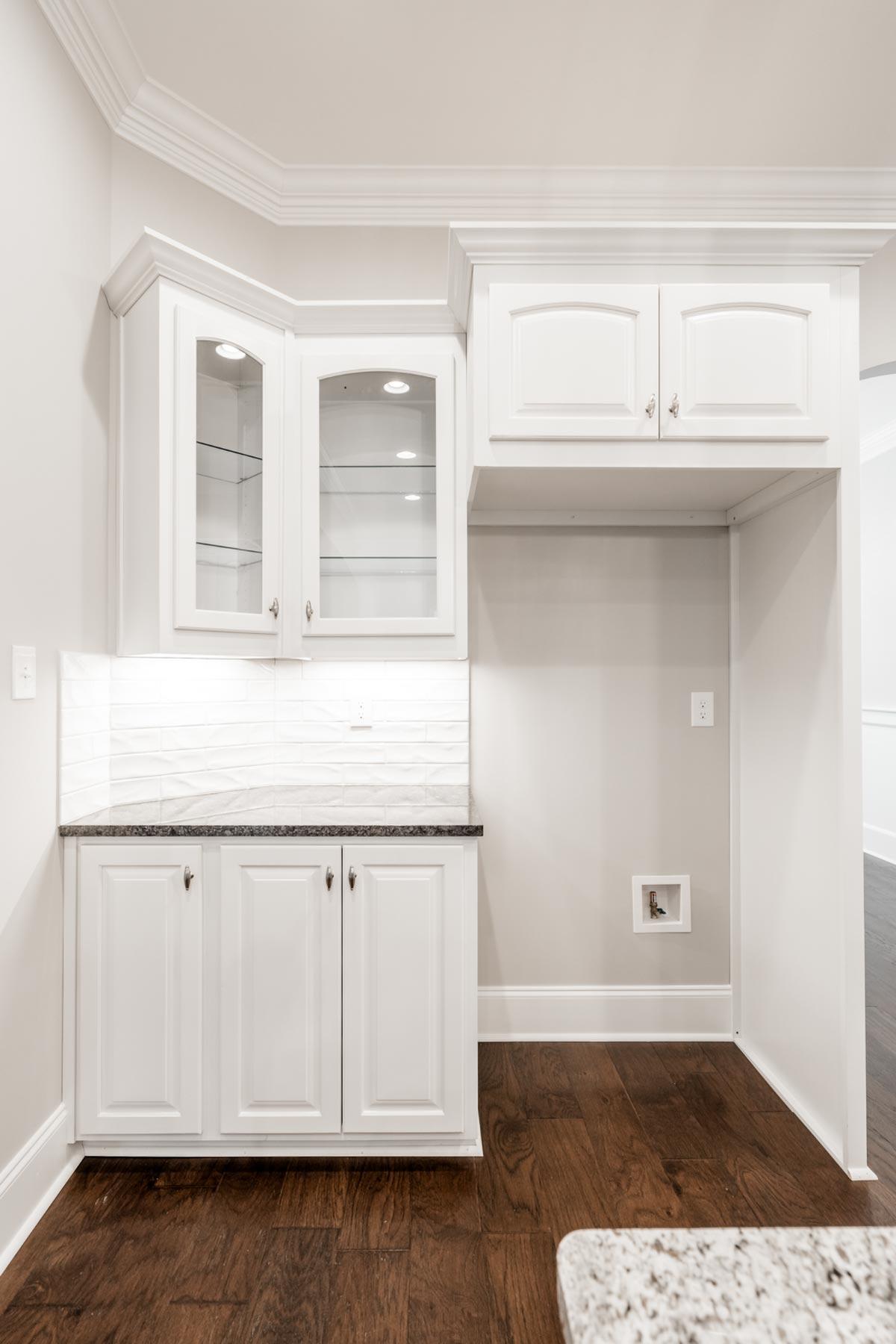 Interior view of a kitchen featuring white cabinetry, glass-front upper cabinets, subway tile backsplash, and a built-in refrigerator space.