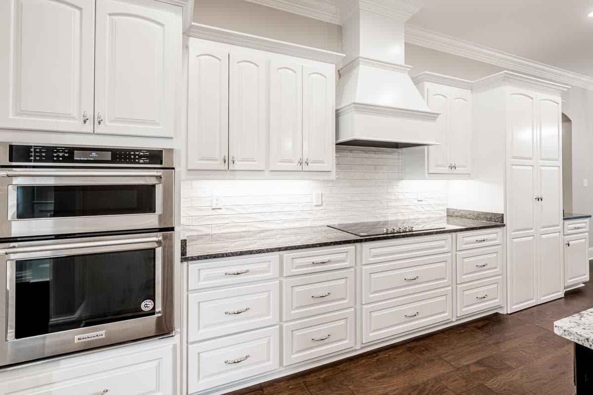 Kitchen interior with white cabinets, dual ovens, subway tile backsplash, cooktop, and granite countertops.