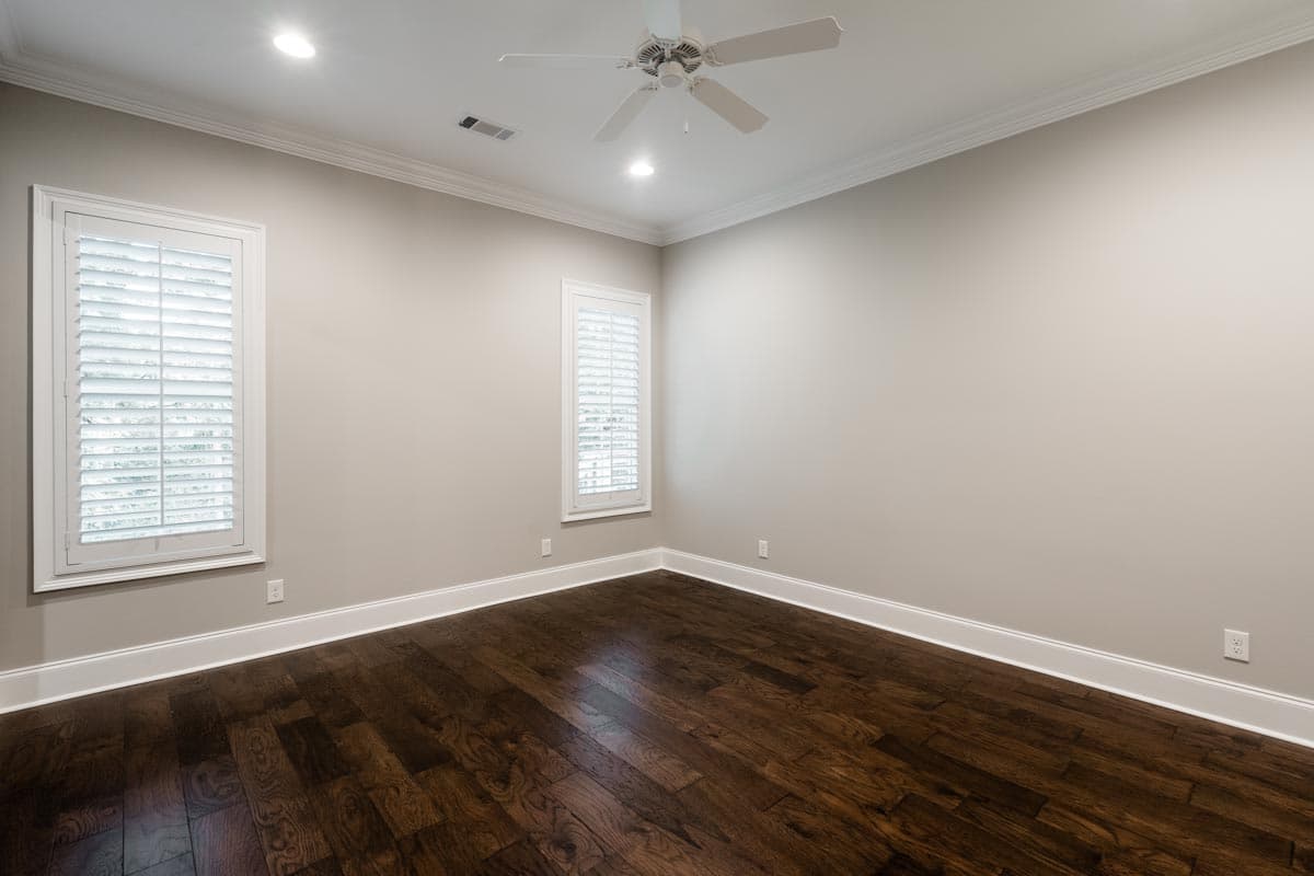 Interior view of an empty room with hardwood floors, two shuttered windows, crown molding, and a ceiling fan.