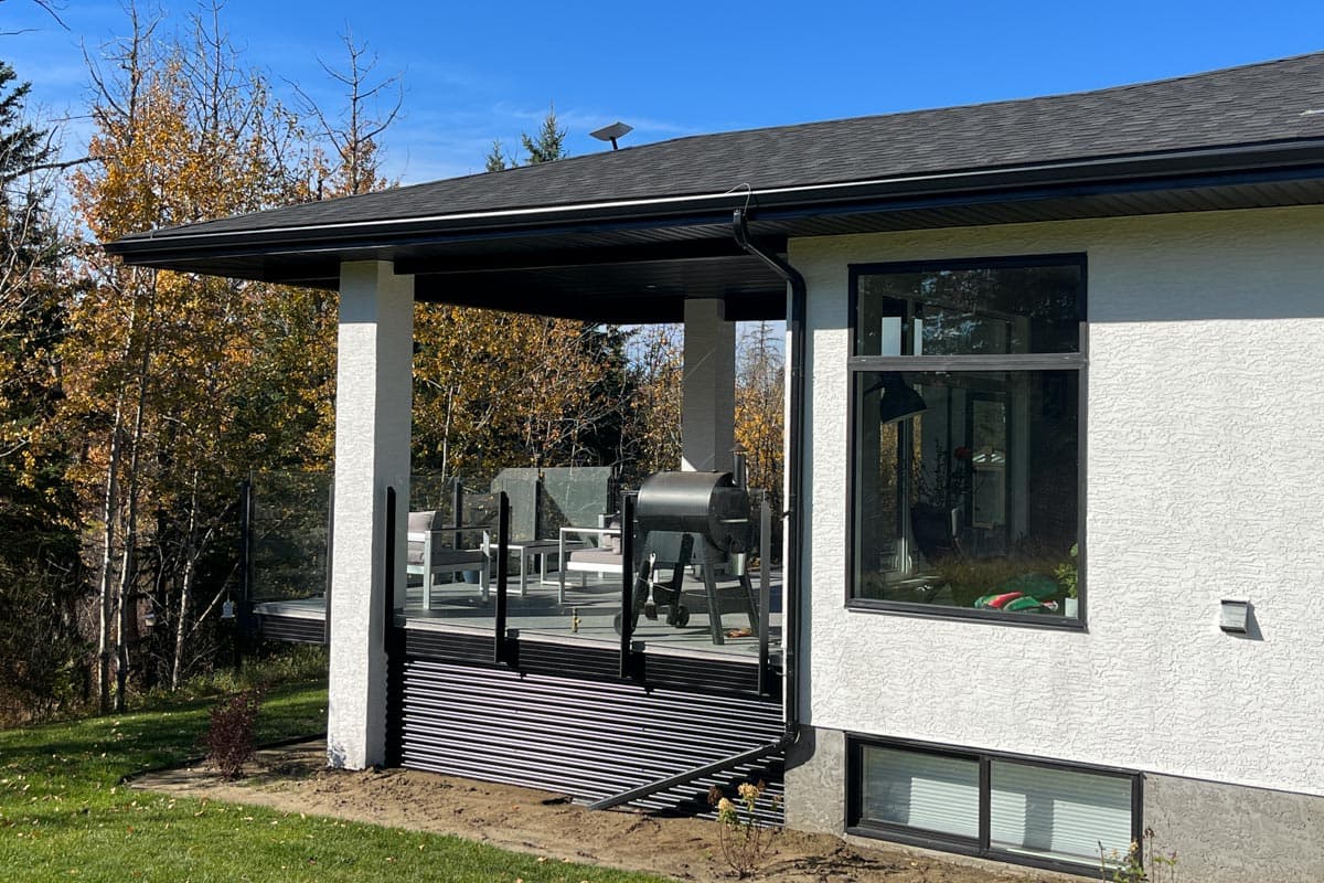 Modern house exterior with covered deck, glass railings, and large windows. Two stories with a dark roof.
