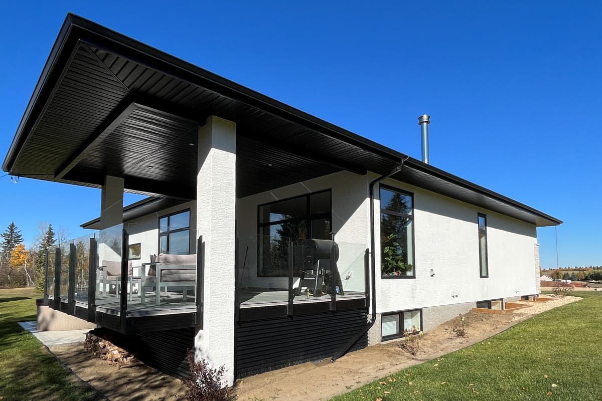Modern house exterior with a covered deck featuring glass railings, large windows, and a broad, dark-paneled soffit under a high-pitched roof.