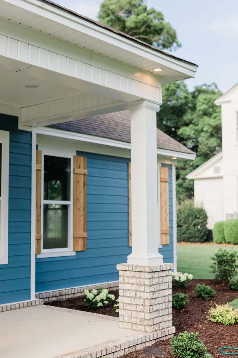 House plan exterior with blue horizontal siding, white trim, covered porch with brick pillar, and wooden shutters.