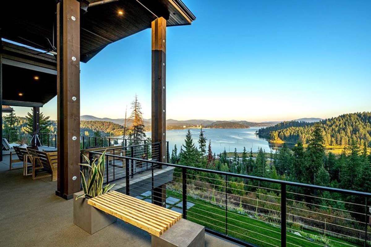 Modern house exterior with expansive deck overlooking a lake and forested hills. Features wood beams and metal railings.