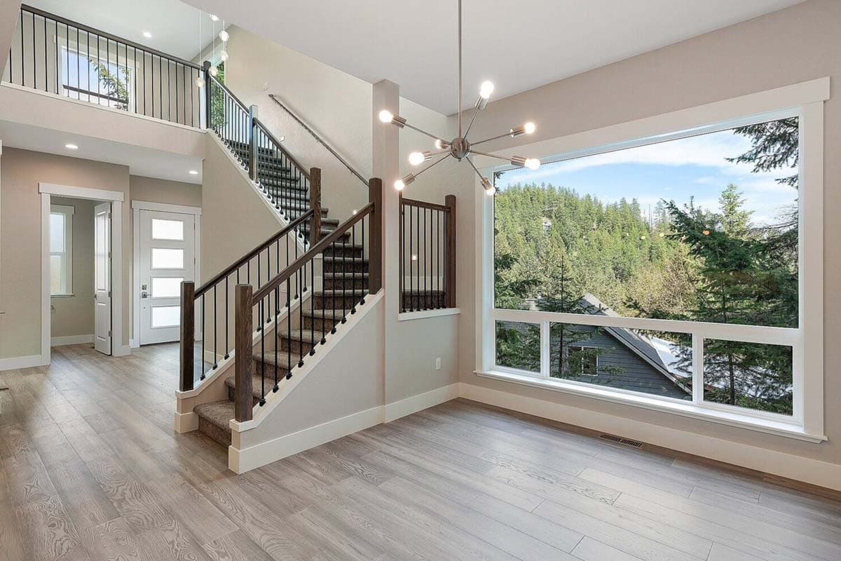 Interior view of a two-story home entrance with a staircase, balcony, and large picture windows overlooking a wooded landscape.