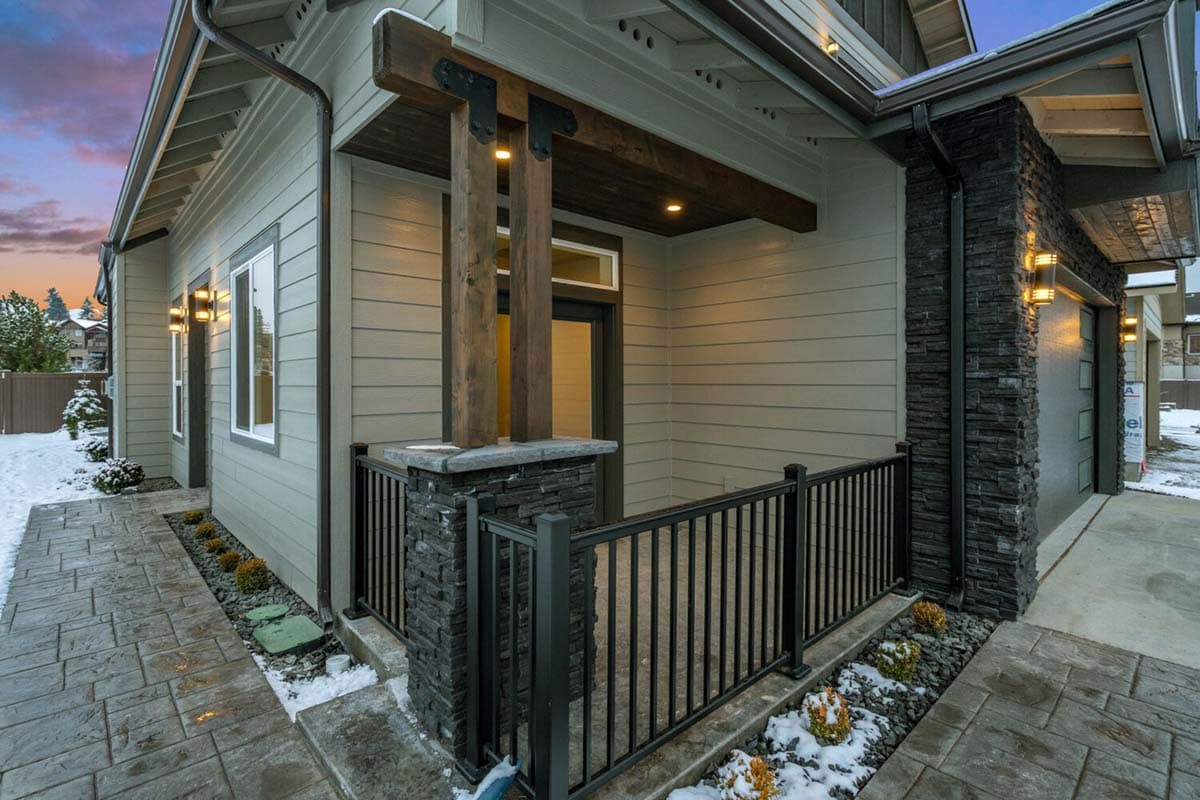 House plan exterior with neutral siding, stone accents on porch column and garage, and exposed wooden beams supporting the roof.