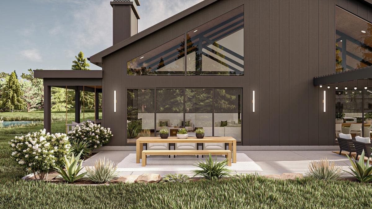 Modern house with a large glass facade. A wooden dining table sits on a patio. White flowers, greenery, and a grassy lawn surround the house.